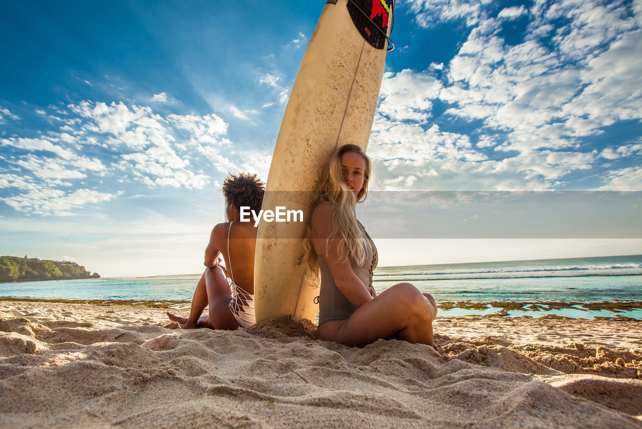 Friends with surfboard sitting on sand at beach against sky