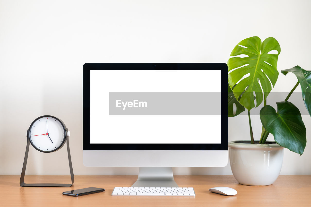 Blank screen of all in one computer, keyboard, mouse, monstera plant pot and clock on wooden table