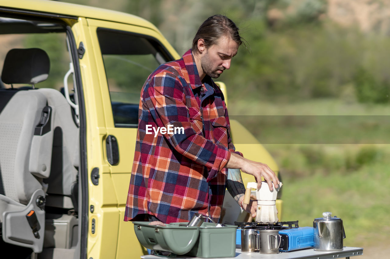 Male traveler in casual clothes assembling moka pot on gas stove while preparing coffee for breakfast outside yellow van during road trip in countryside in morning