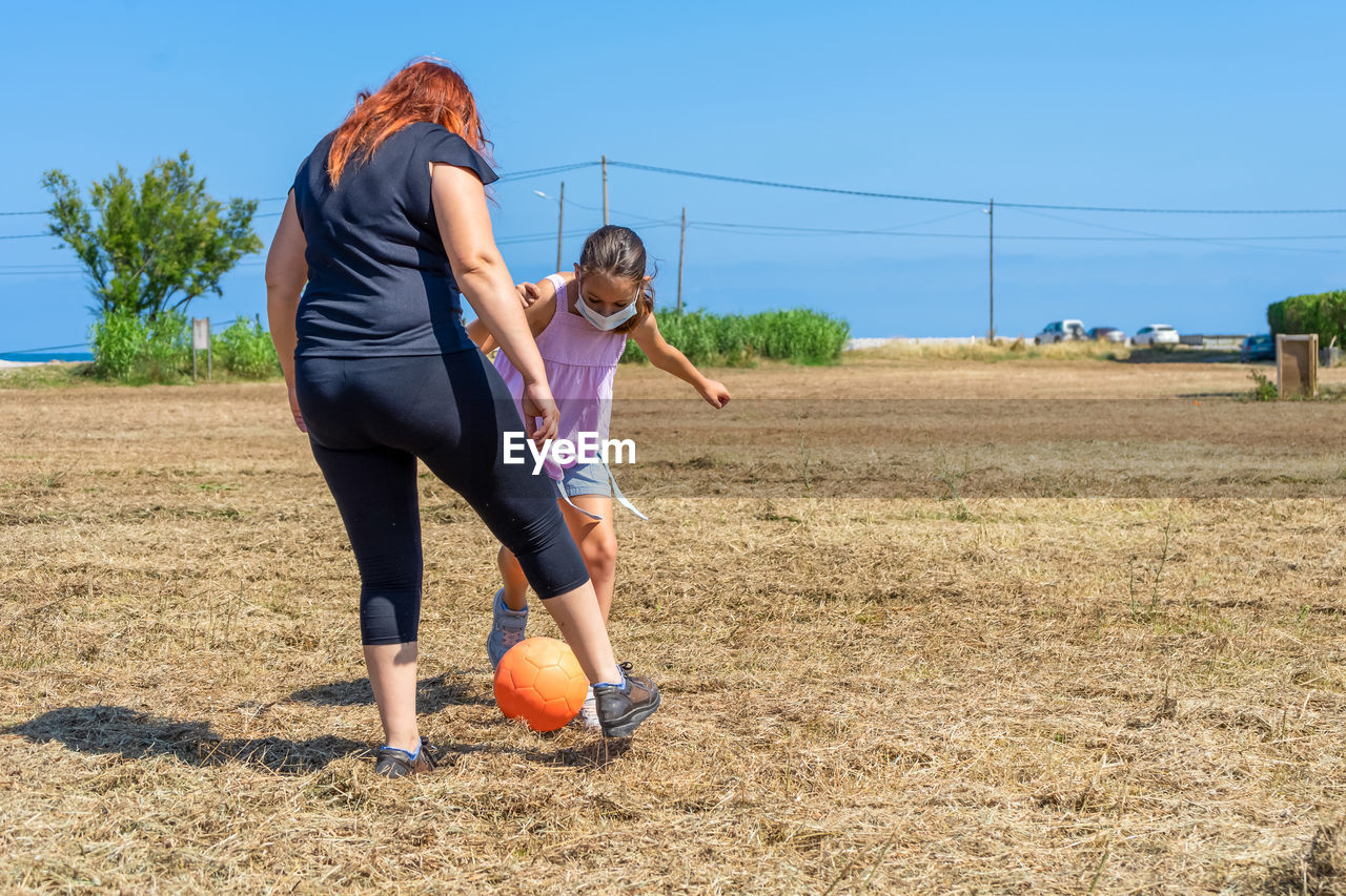 Full length of mother and daughter with protective face mask playing football on field