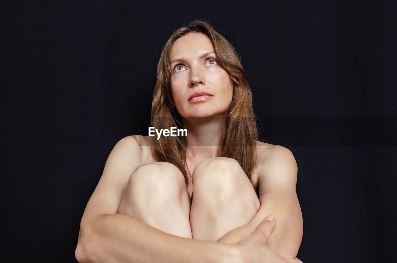 Portrait of woman on black background, sitting, hugging knees.