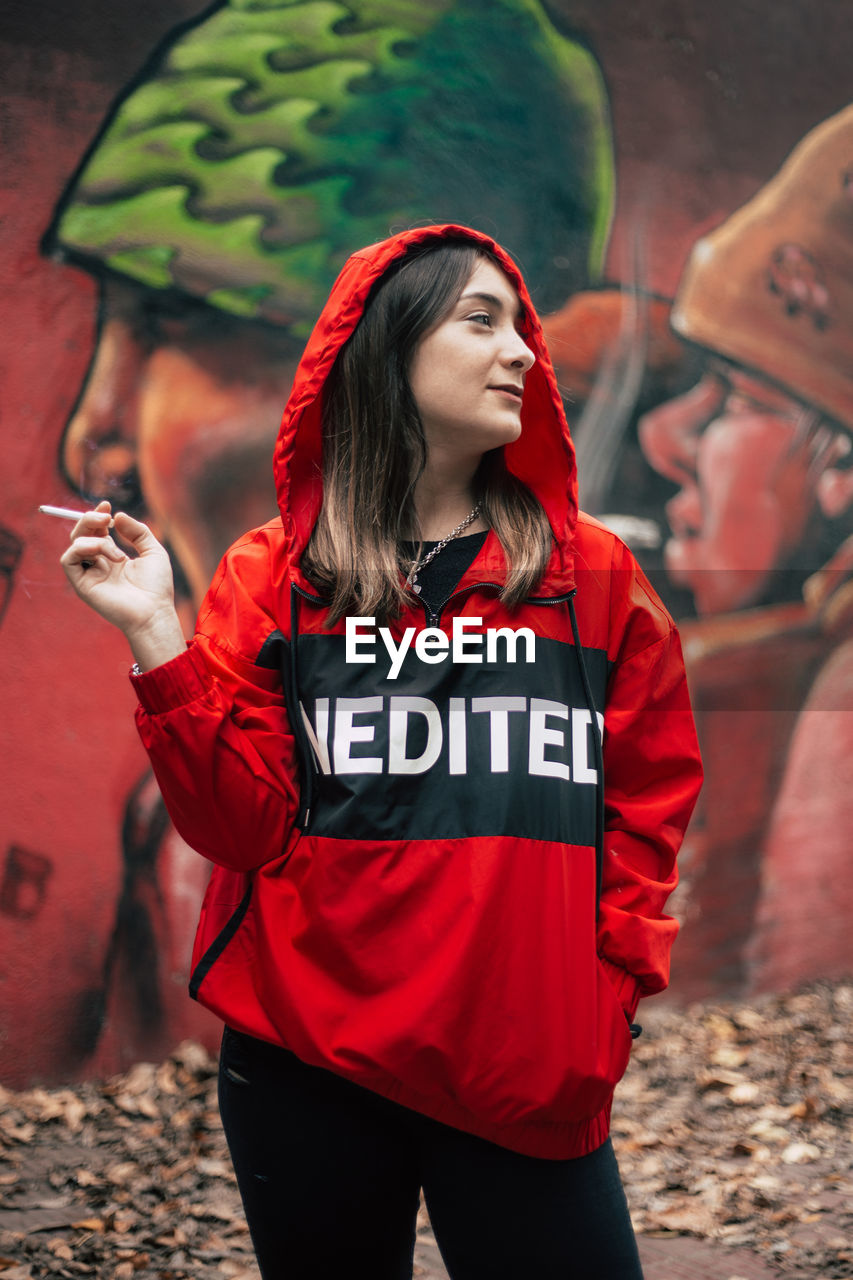 Woman in red hood clothing smoking cigarette against graffiti wall