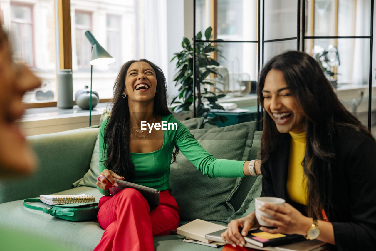 Cheerful businesswoman laughing while sitting with female colleague on sofa at office