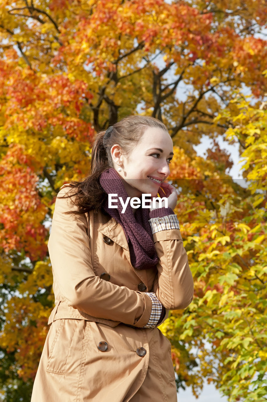 Smiling woman standing against trees in park during autumn