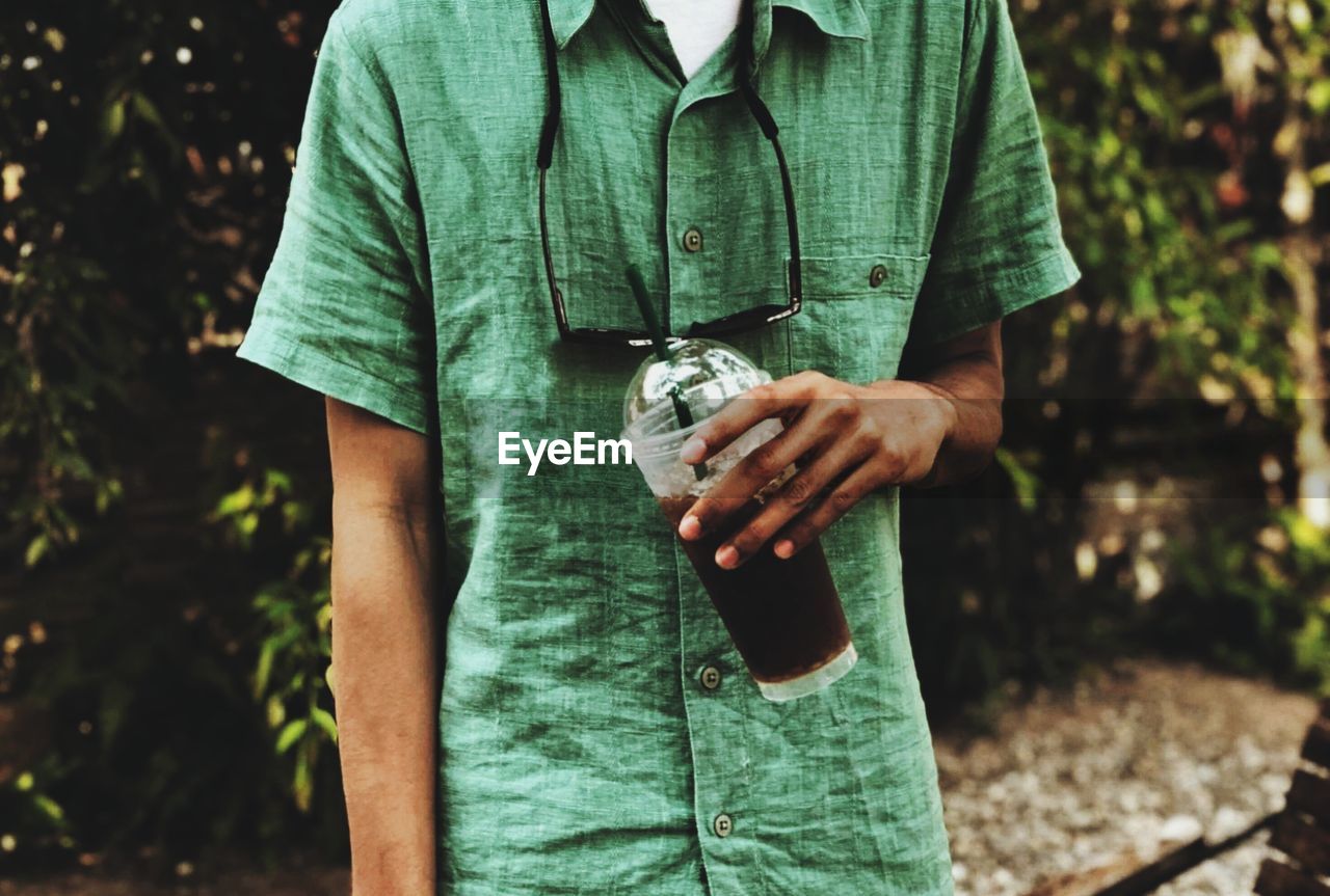 Midsection of man holding disposable glass with drink against trees