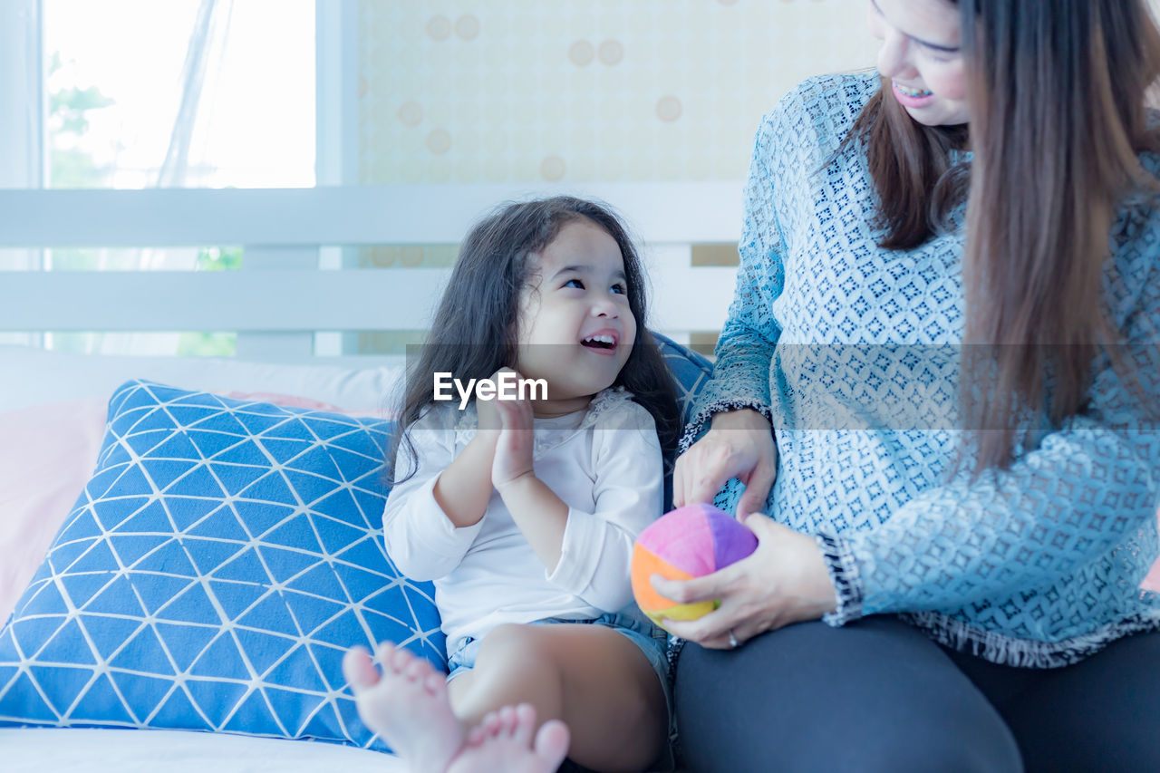 Pregnant mother holding ball by daughter while sitting on bed at home