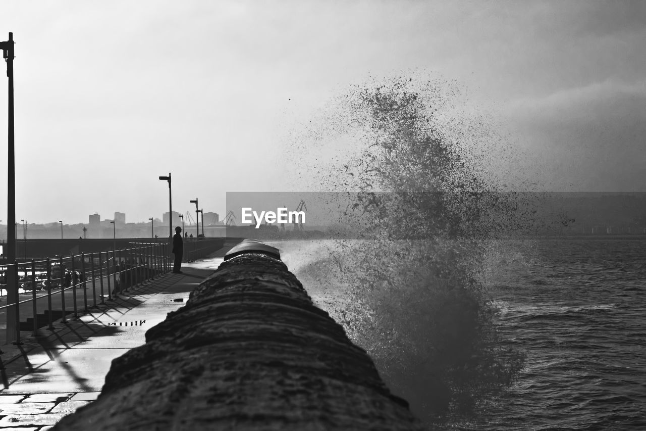 Silhouette person standing by waves splashing on shore against sky