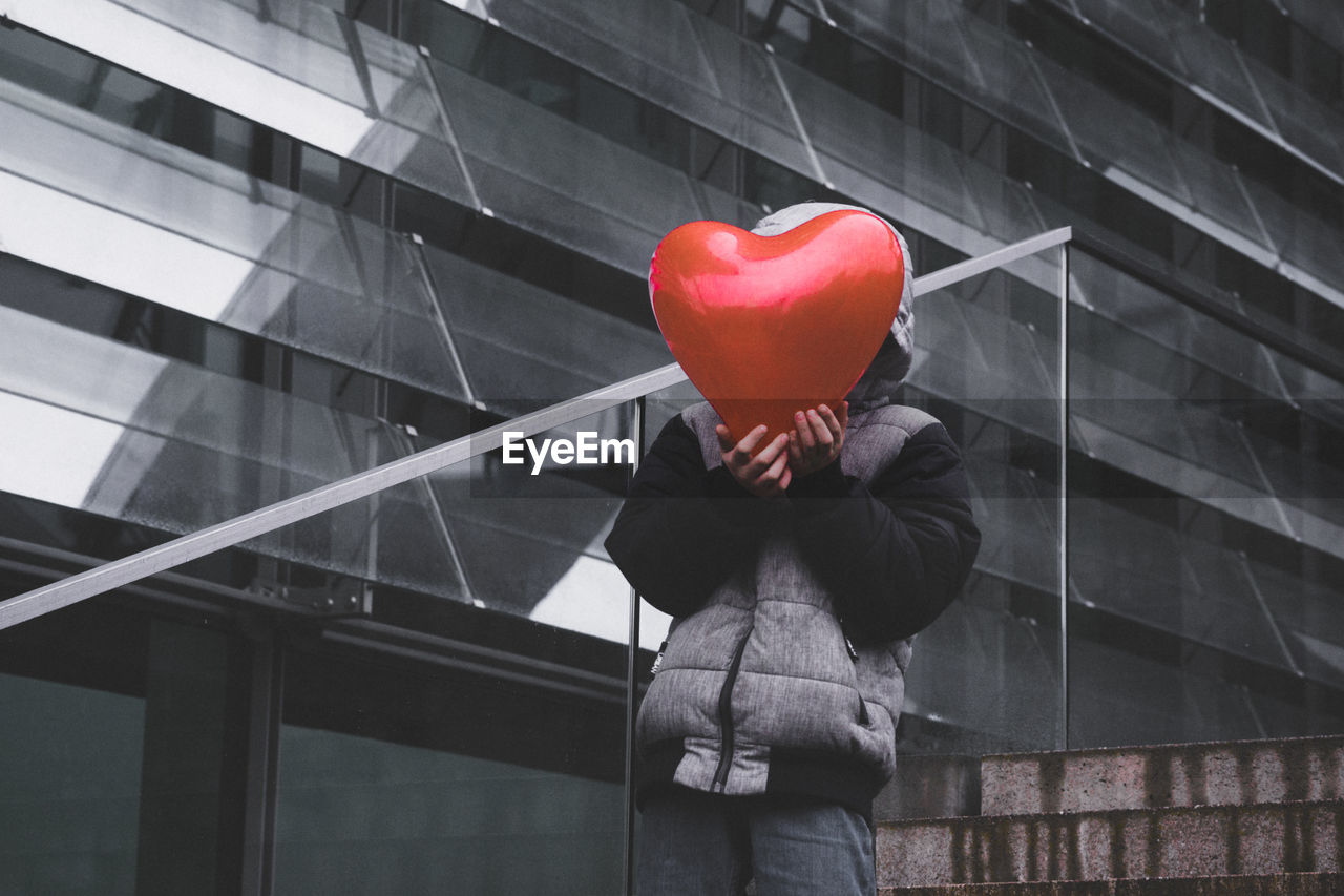 Boy wearing warm clothing while covering face with red heart shape balloon in city