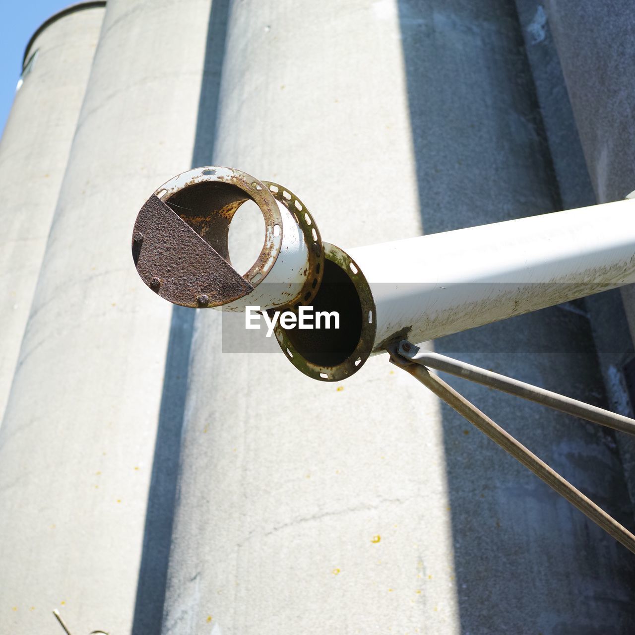 Low angle view of metallic pipe against silos during sunny day