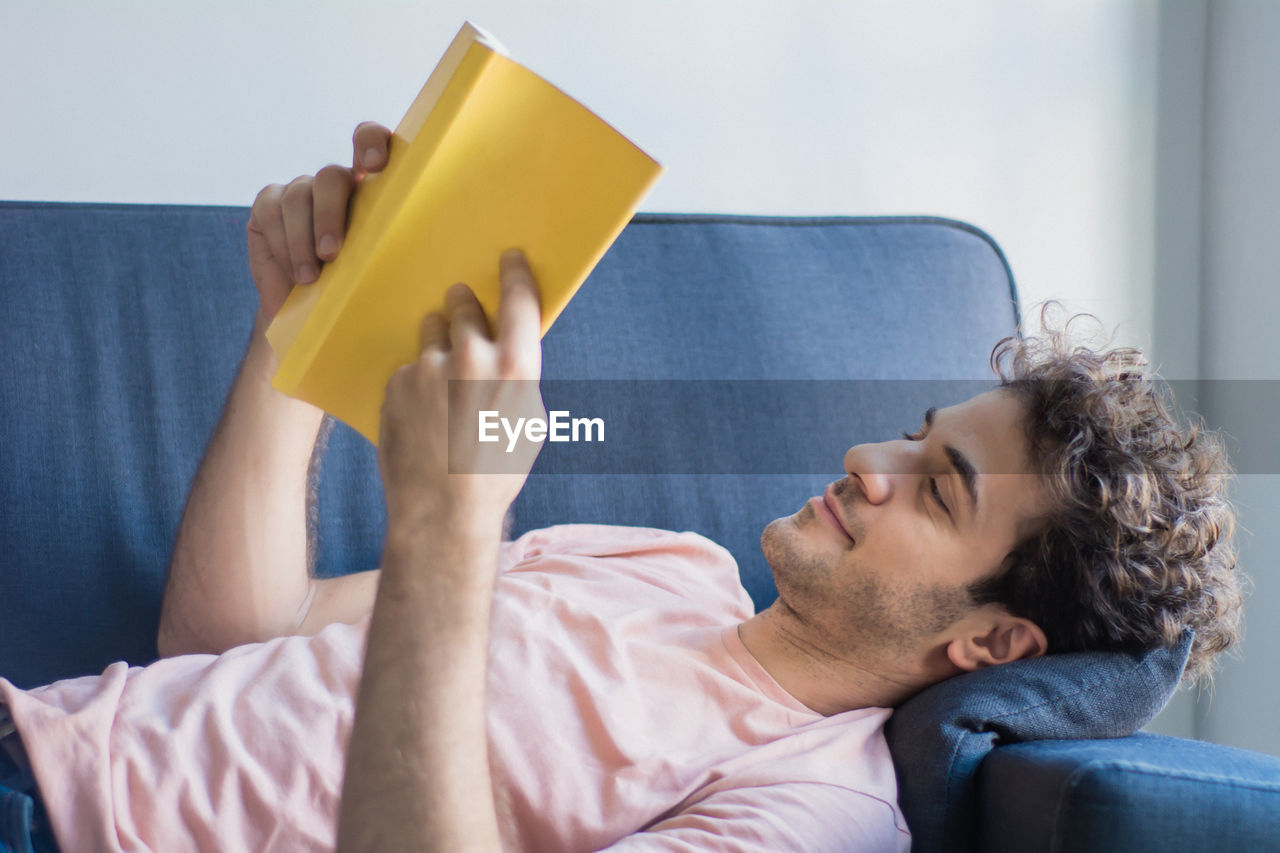 MIDSECTION OF MAN READING BOOK AGAINST WALL AT HOME