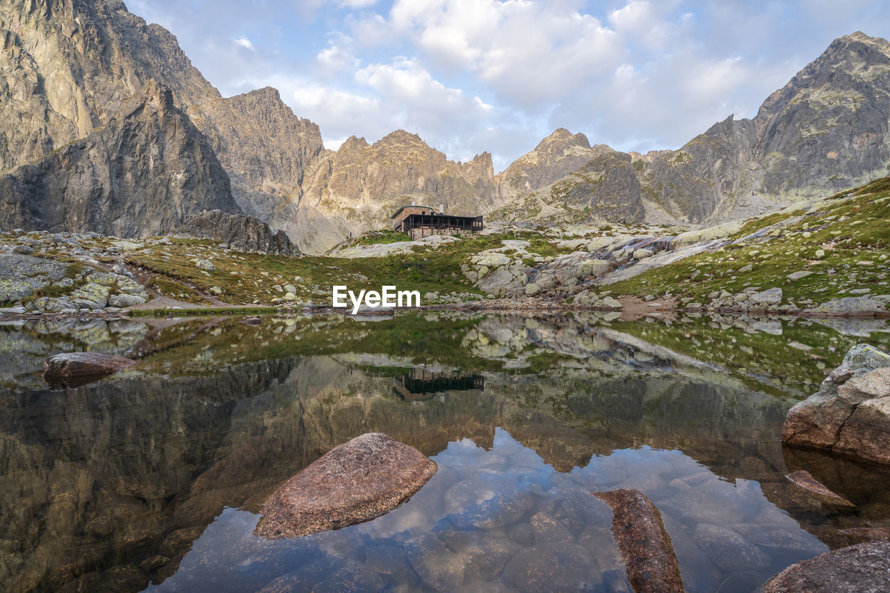 Alpine hut surrounded by mountains reflected in foreground tarn during sunrise, slovakia, europe