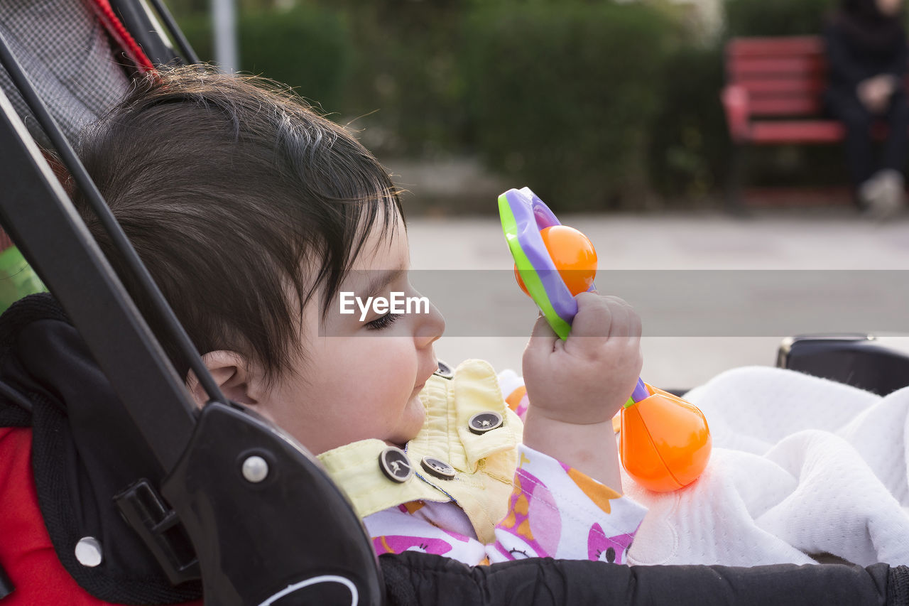 Side view of cute baby girl holding toy while lying on stroller