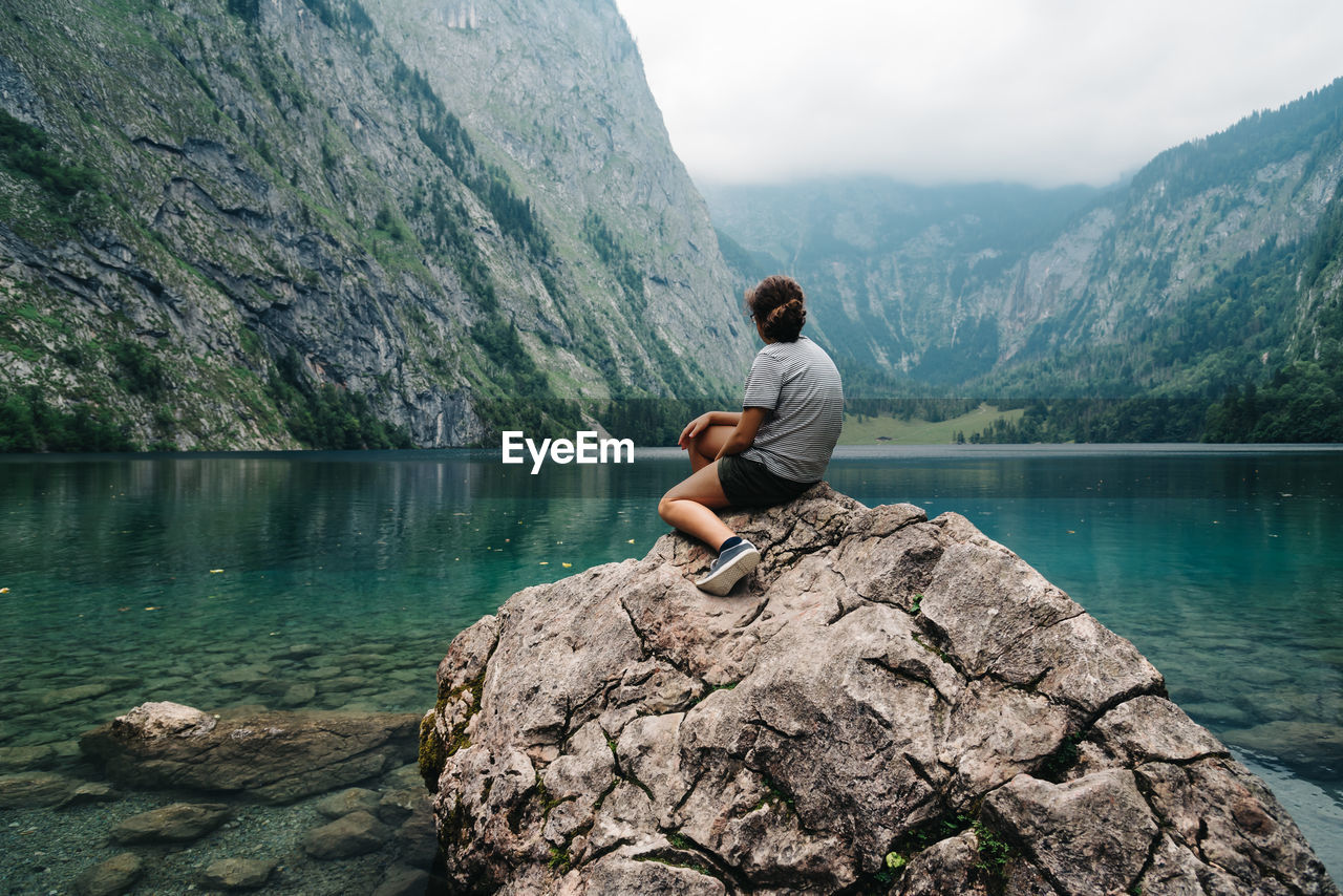 Rear view of young woman sitting at lake against mountain