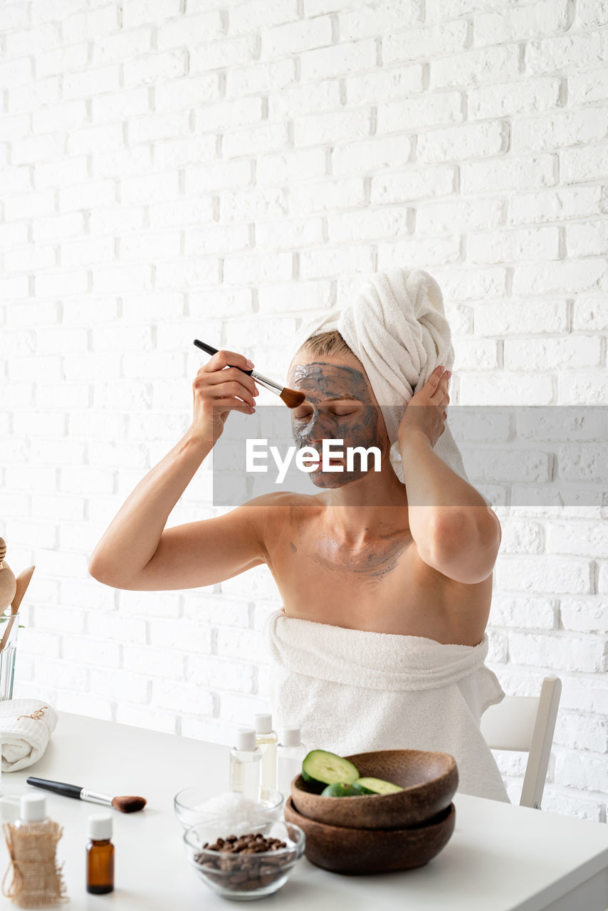 Young woman wearing white towels applying spa facial mask on her face with a cosmetic brush