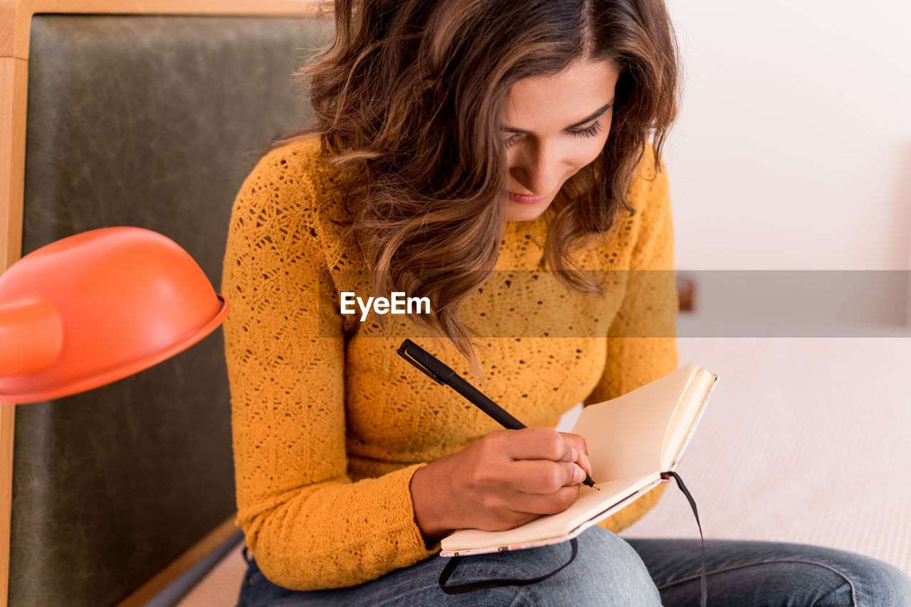 Smiling woman writing in diary while sitting on bed at home