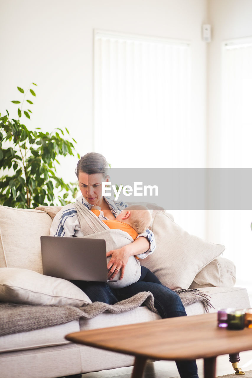 Multi-tasking mother using laptop while caring baby girl on sofa at home office