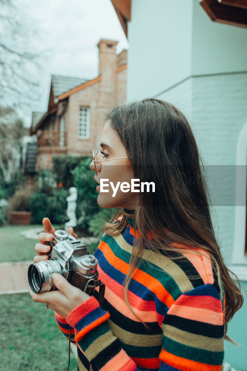 PORTRAIT OF YOUNG WOMAN PHOTOGRAPHING AGAINST BUILDING