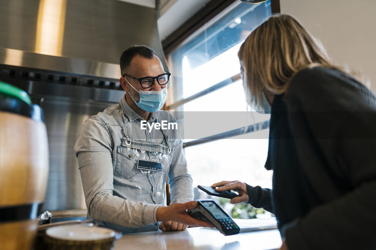 Female customer paying through smart phone while looking at waiter in a bar during pandemic