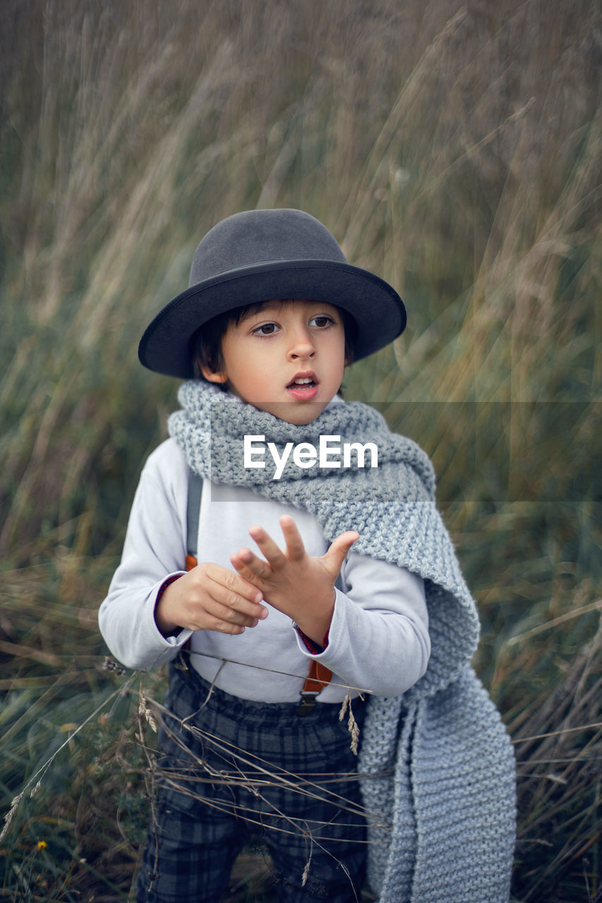 Boy child in plaid pants, hat, suspenders and scarf stands in a field in autumn