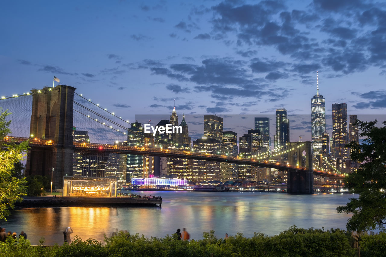 Illuminated brooklyn bridge over river against buildings at night