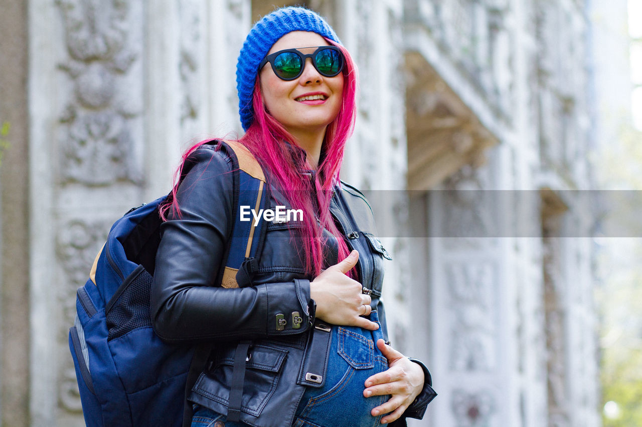adult, one person, fashion, smiling, women, happiness, architecture, clothing, sunglasses, glasses, blue, young adult, person, city, lifestyles, emotion, portrait, jacket, standing, leisure activity, backpack, cheerful, day, outdoors, city life, spring, waist up, enjoyment, female, technology, copy space, nature, arts culture and entertainment, human face, travel, goggles, smile, teeth, hat, casual clothing, travel destinations, warm clothing, built structure, focus on foreground, tourism, building exterior, carefree, positive emotion, hairstyle