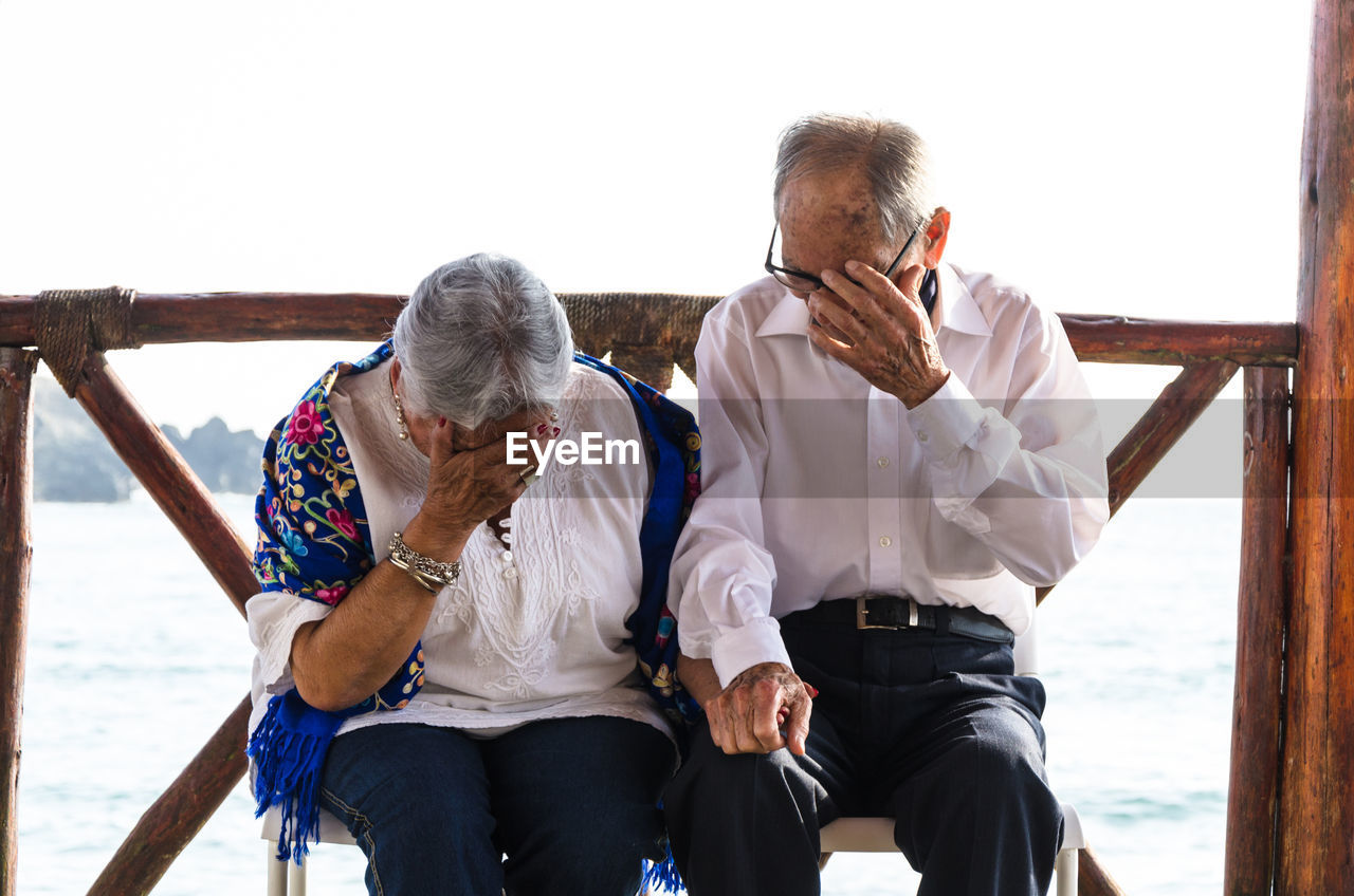 Senior couple covering face while sitting on bench with sea in background