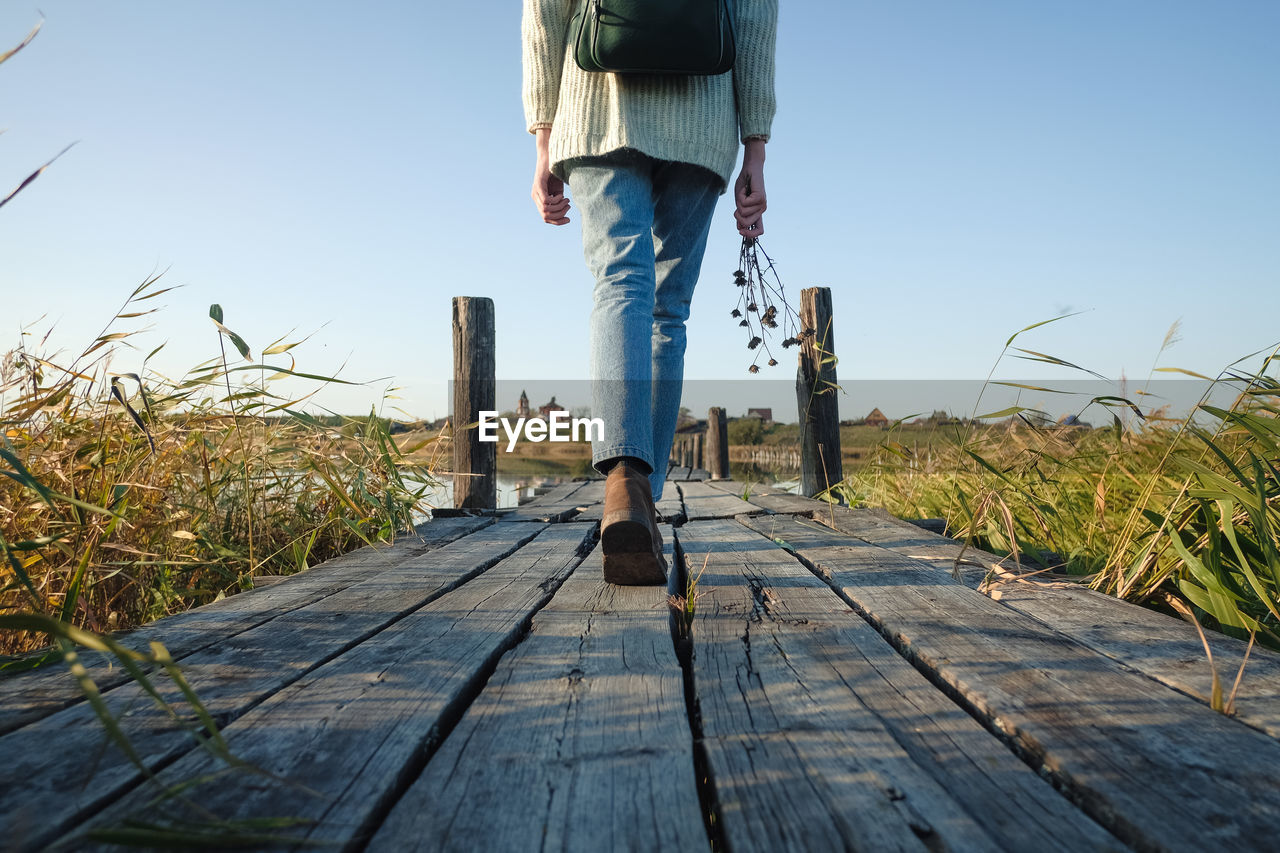 LOW SECTION OF MAN STANDING ON BOARDWALK AGAINST CLEAR SKY