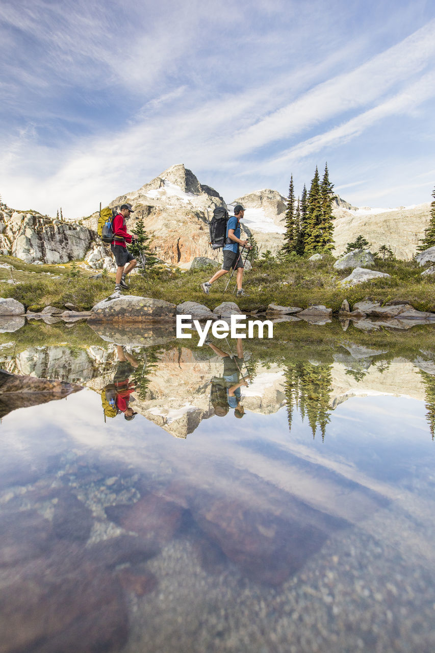 Side view and reflection of backpackers hiking beside alpine lake.