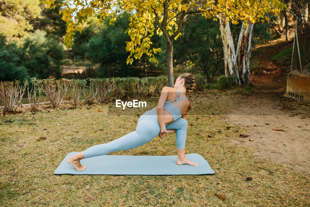 Full body side view of flexible slim female standing on mat and performing bound revolved crescent lunge pose while practicing yoga in park