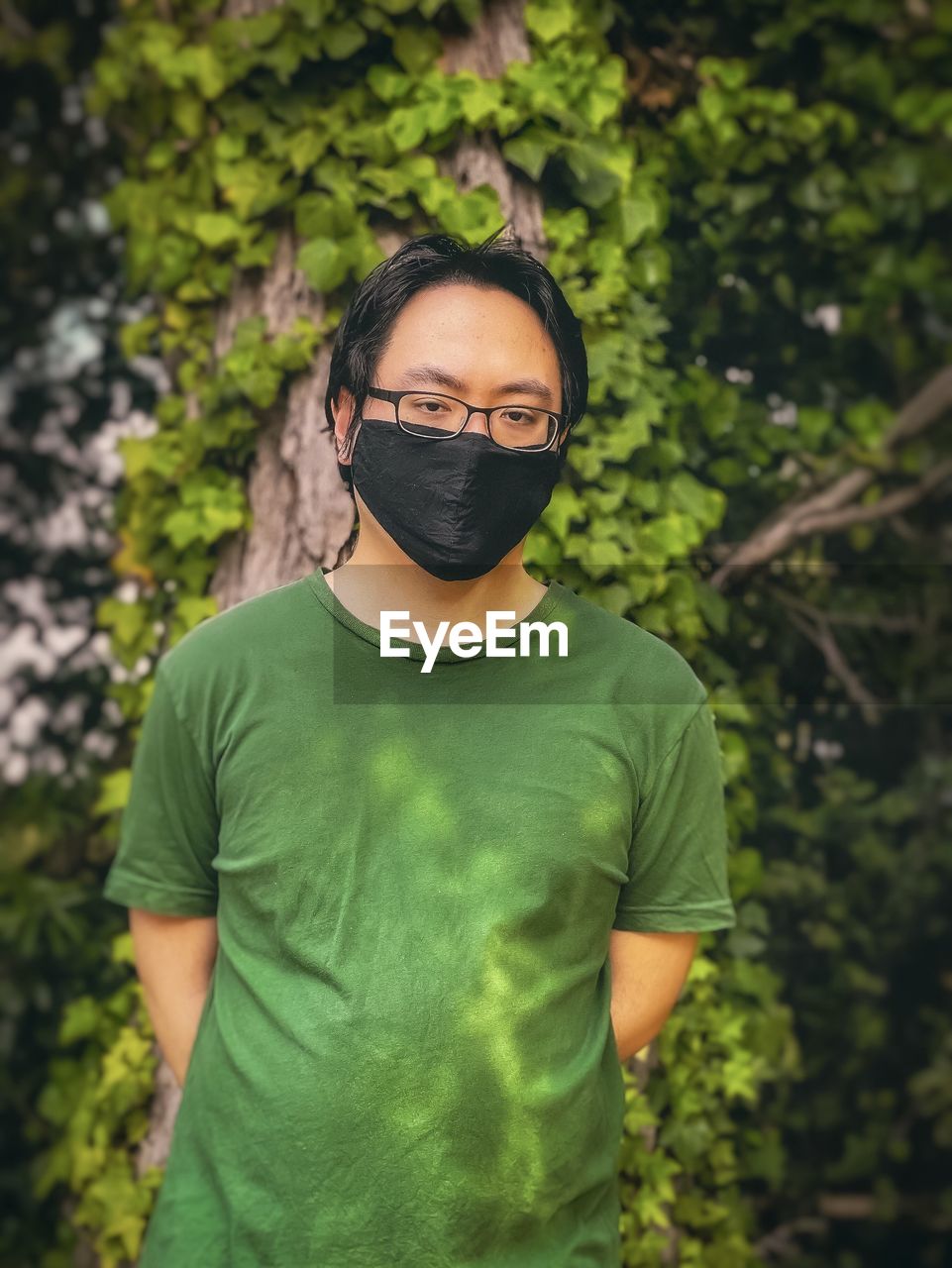 Young man in eyeglasses and mask standing against trees and plants.