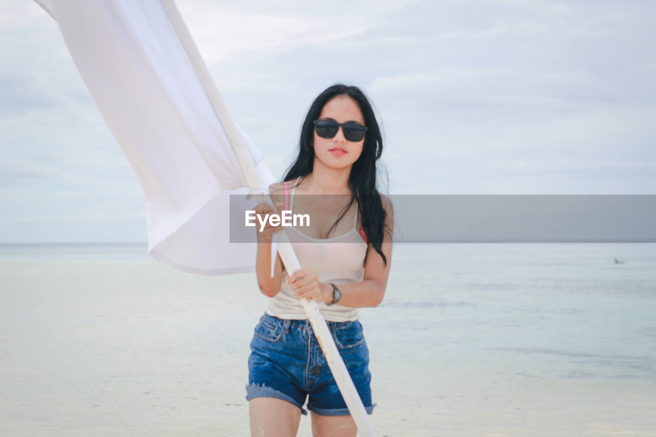 YOUNG WOMAN WEARING SUNGLASSES STANDING AT BEACH