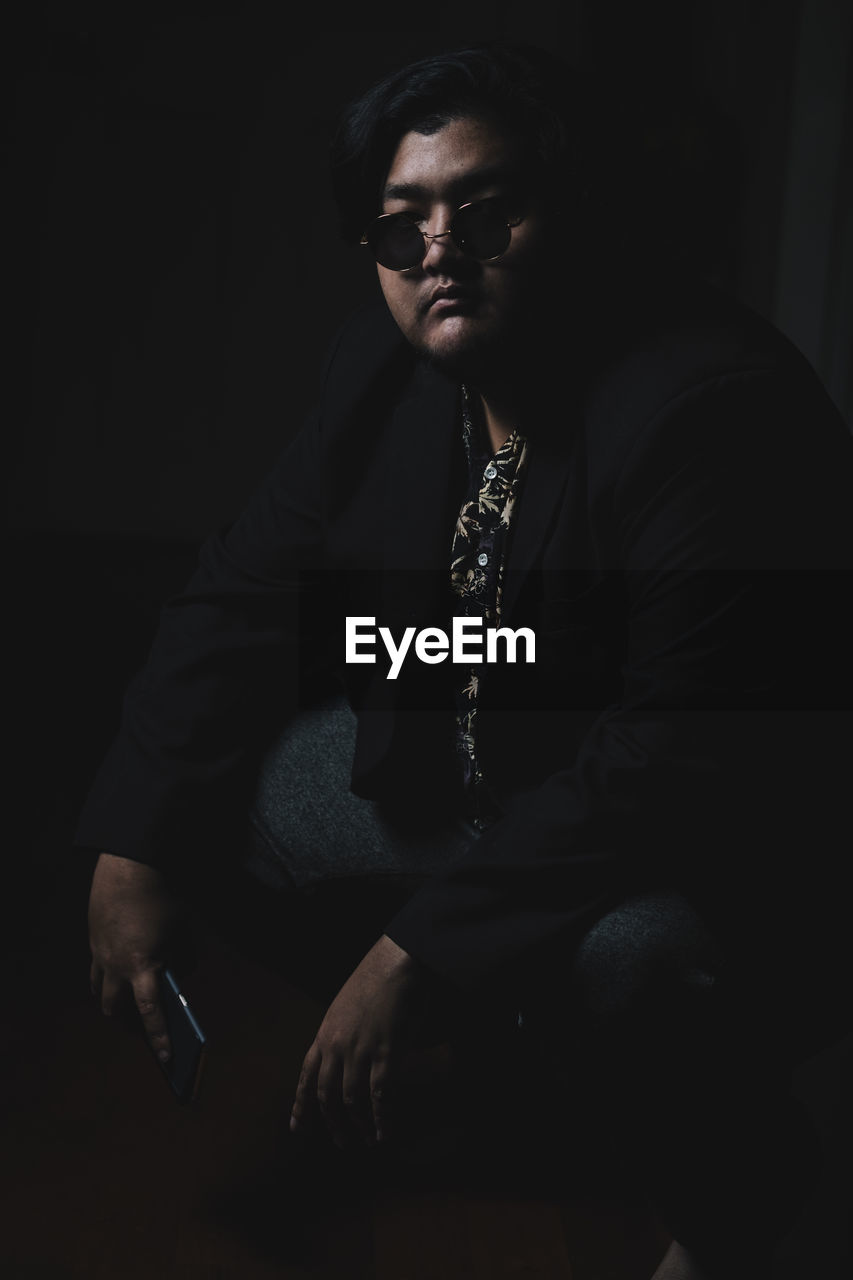 Young man holding mobile phone while sitting against black background