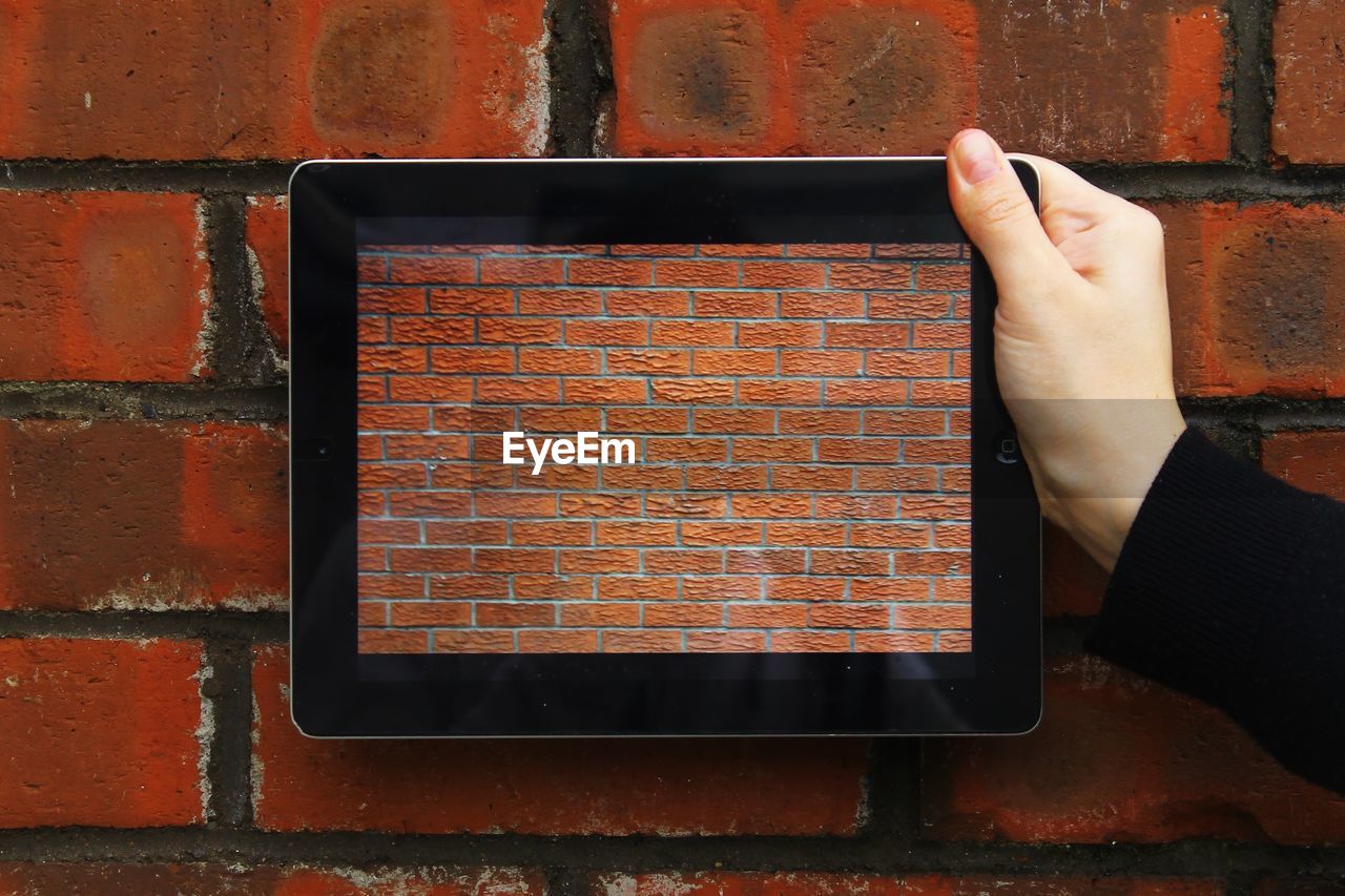 Cropped image of hand holding digital tablet with brick wall picture