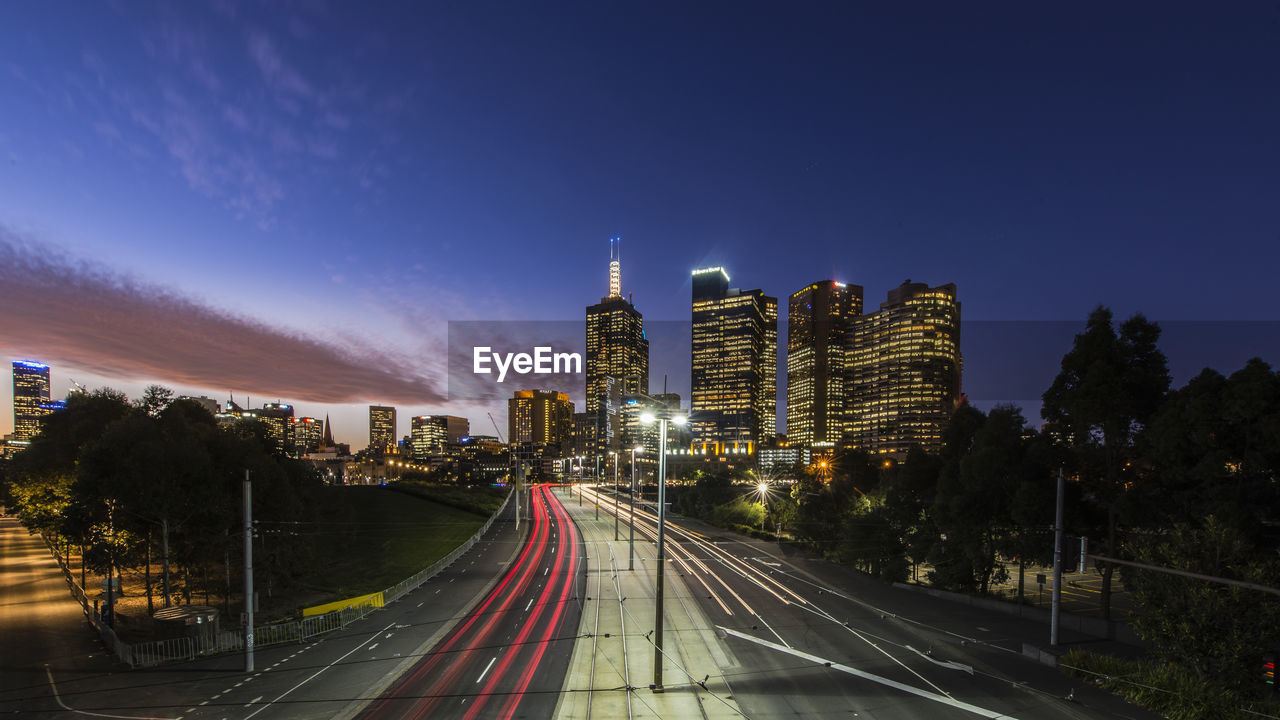 Light trails on road amidst buildings against sky at night