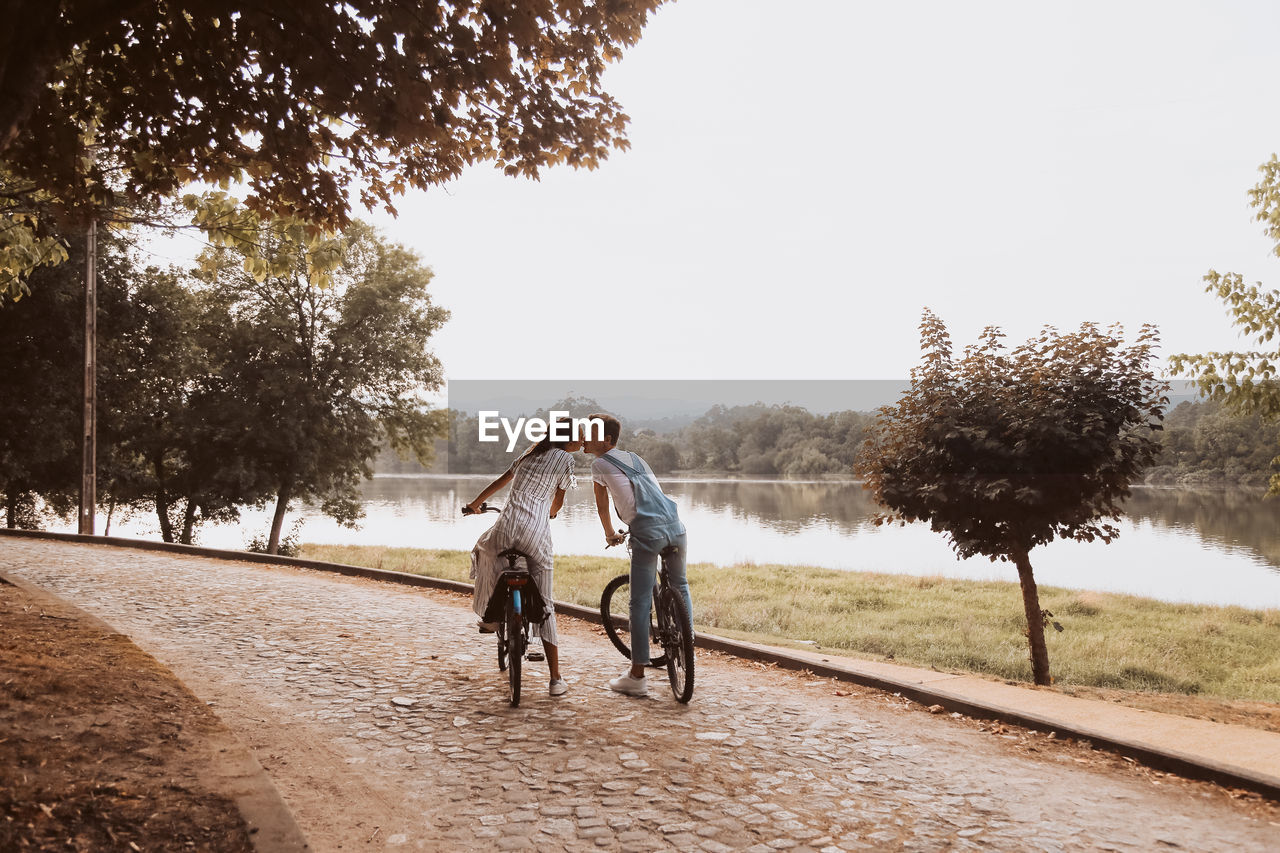 Couple kissing while sitting bicycles at lakeshore against clear sky