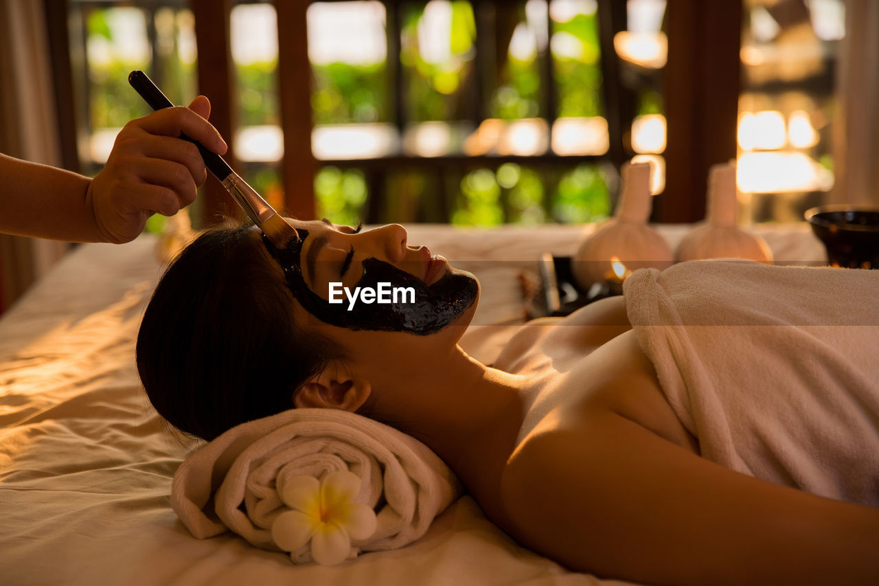 Cropped hand applying facial mask to young woman lying on bed in spa