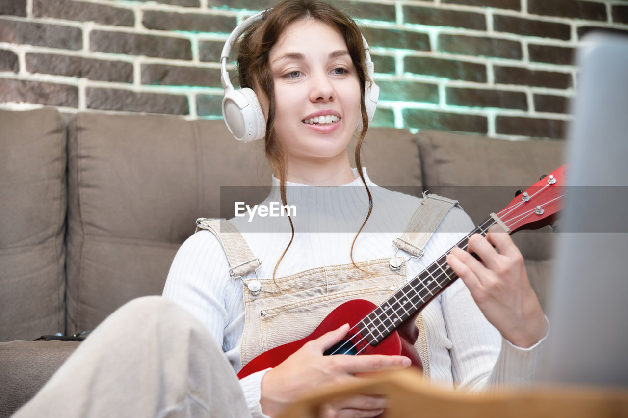 Young cheerful girl in a sweater is learning to play the ukulele. online learning, home education