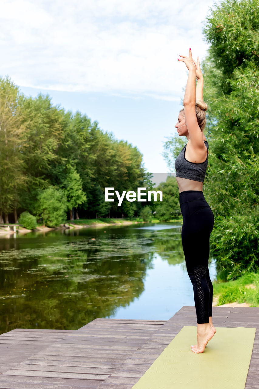 A woman stands on a green rug, by a pond , in summer, doing yoga.