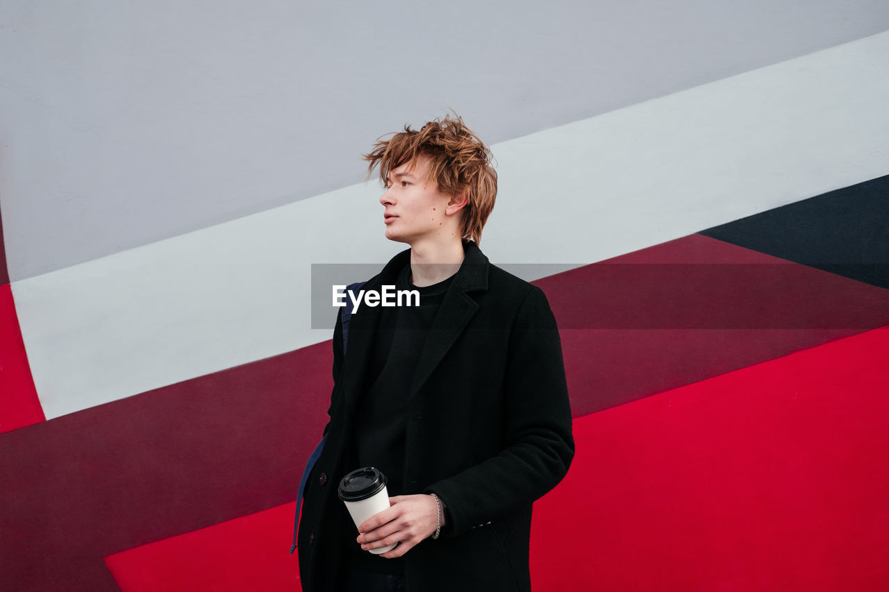 Young man in black coat with cup of coffee in hands looking away outdoors