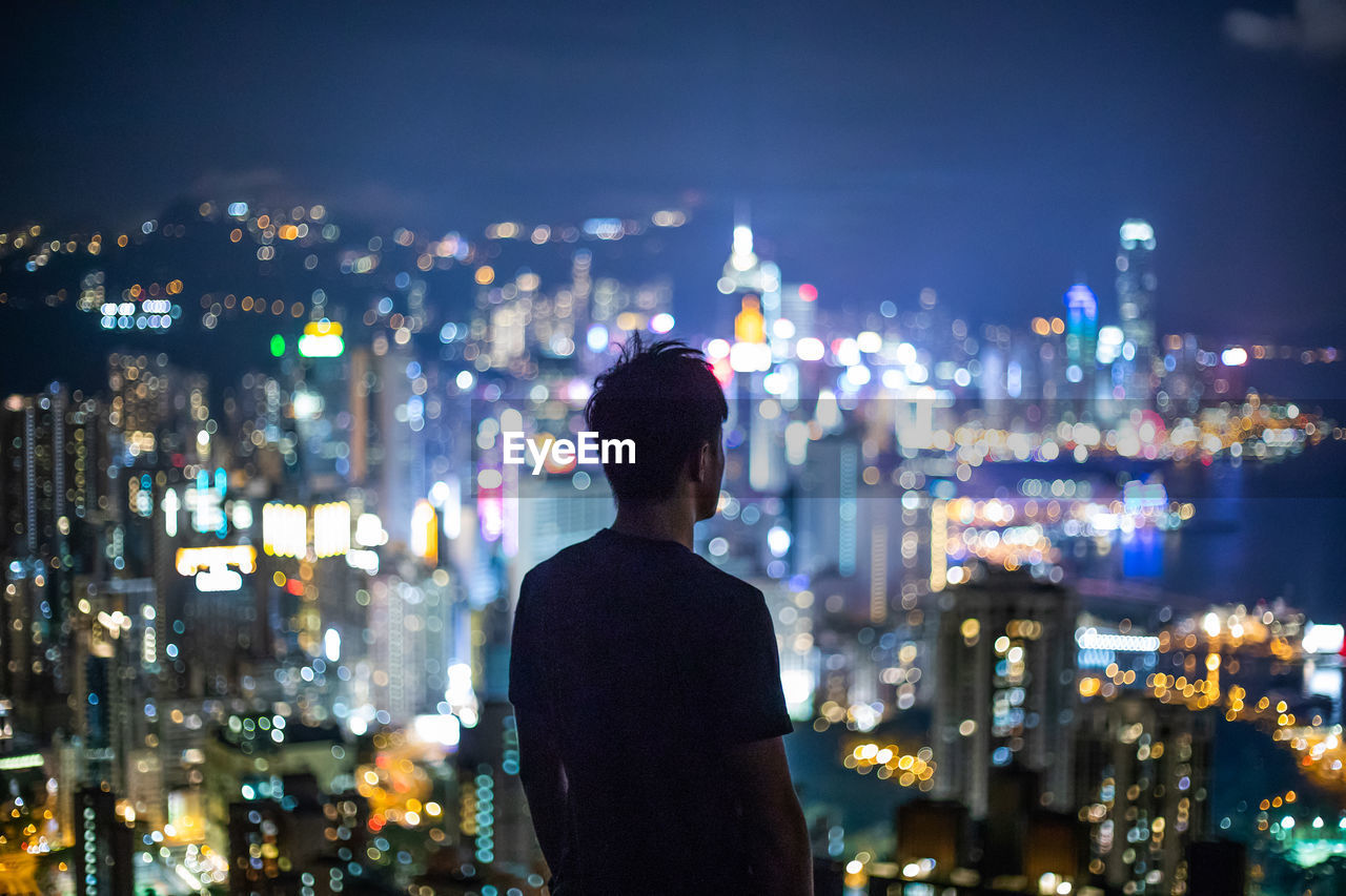Rear view of man looking at illuminated city buildings at night