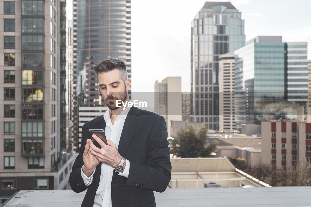 Man using smart phone outside on downtown rooftop with skyscrapers in background.