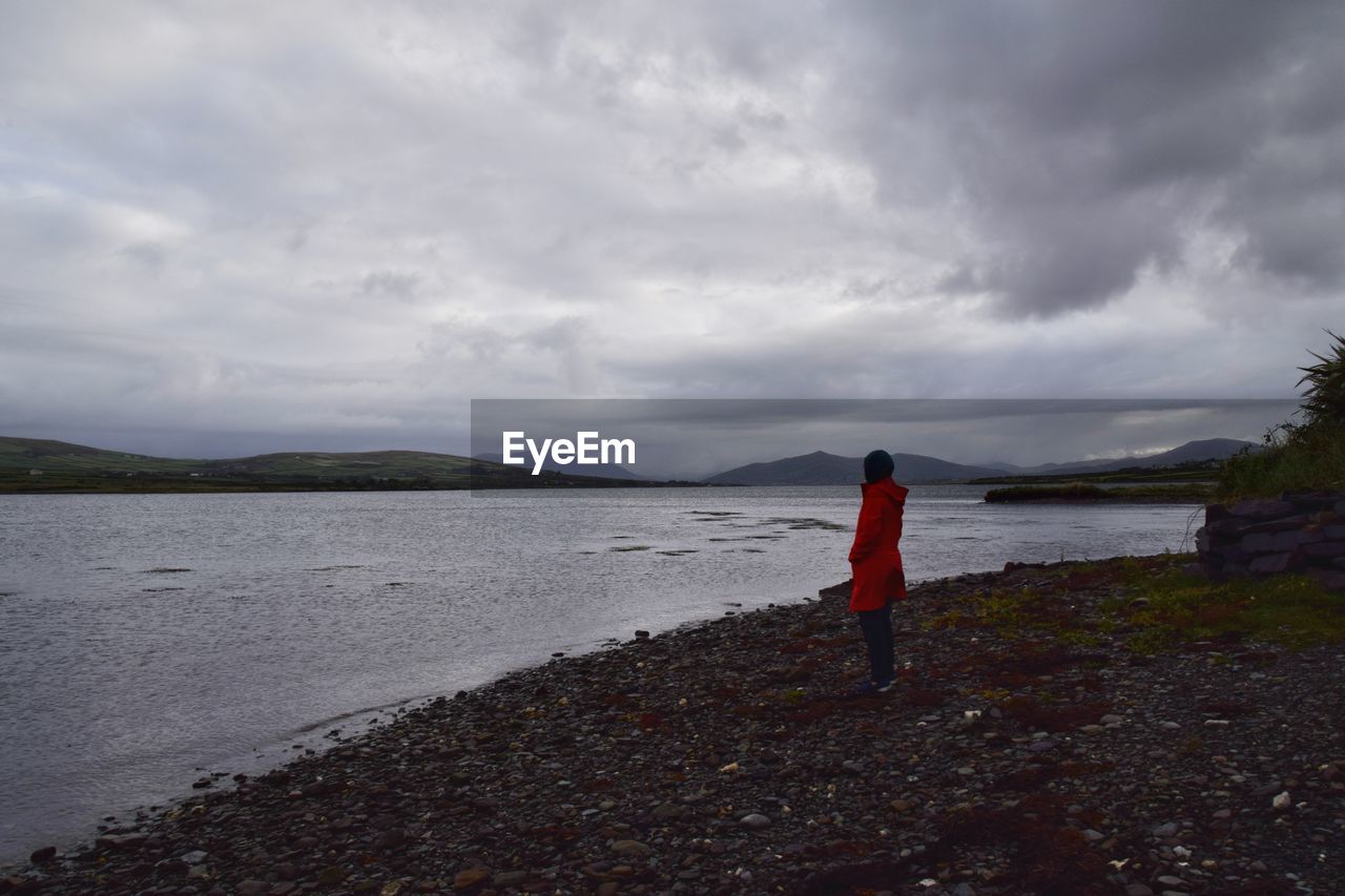 REAR VIEW OF WOMAN STANDING ON SHORE AGAINST SKY