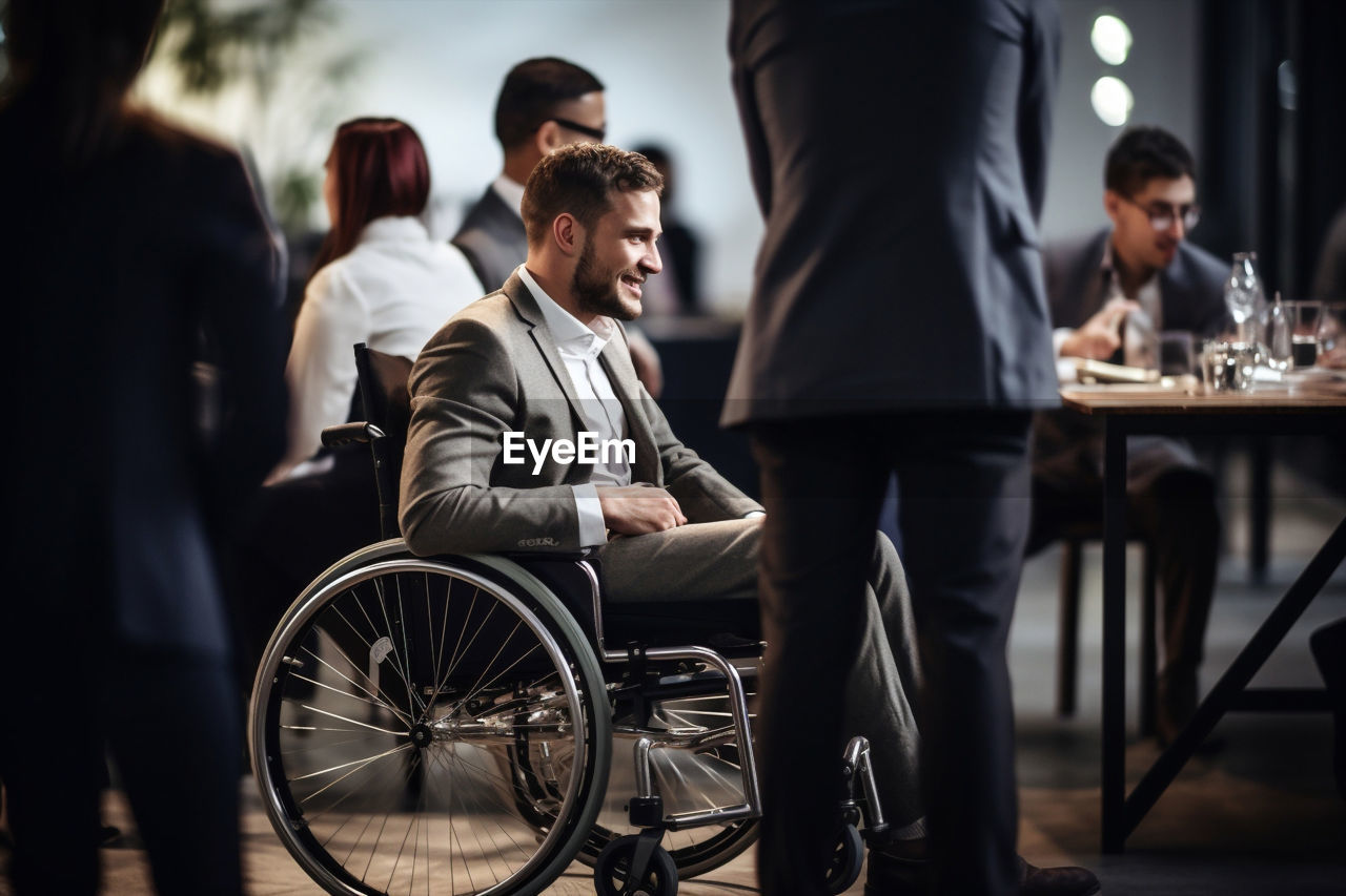 wheelchair, adult, men, group of people, sitting, medical equipment, business, young adult, women, differing abilities, clothing, physical impairment, person, businessman, table, female, communication, indoors, professional occupation, talking, corporate business, city, lifestyles