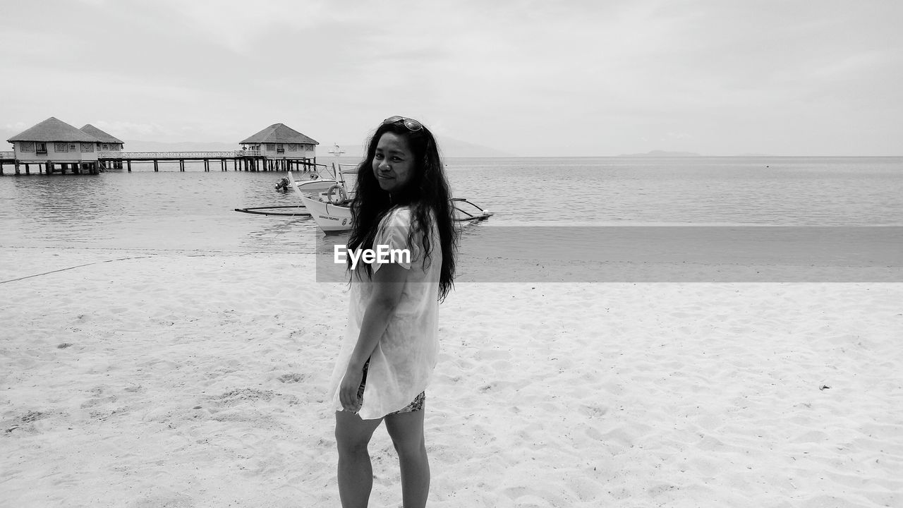 PORTRAIT OF YOUNG WOMAN STANDING ON BEACH AGAINST SEA