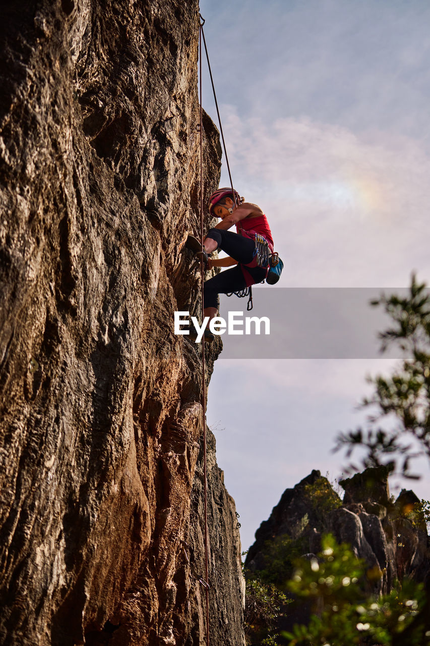 Low angle full length of active senior woman climbing on rocky cliff against sky