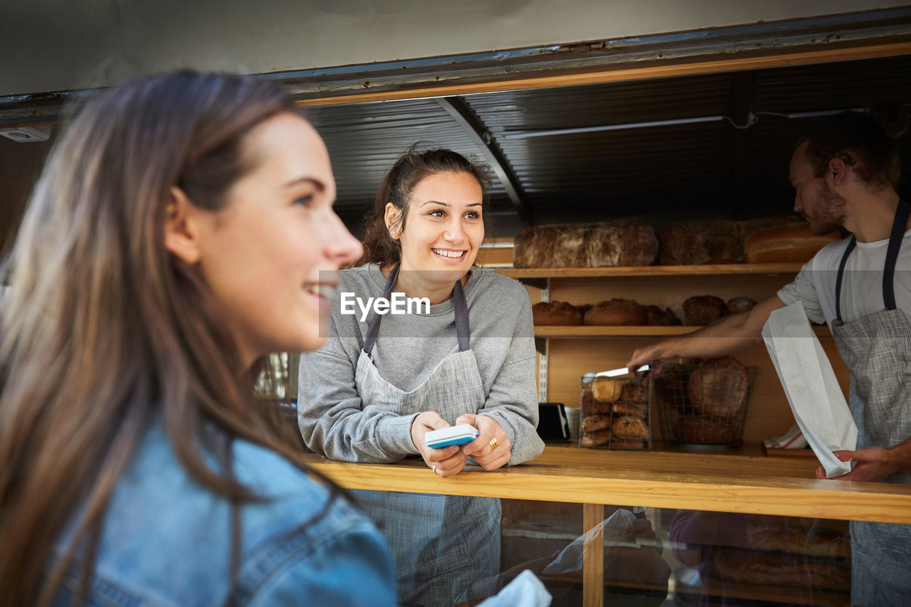 Smiling saleswoman accepting payment from customer while colleague packing bread at concession stand