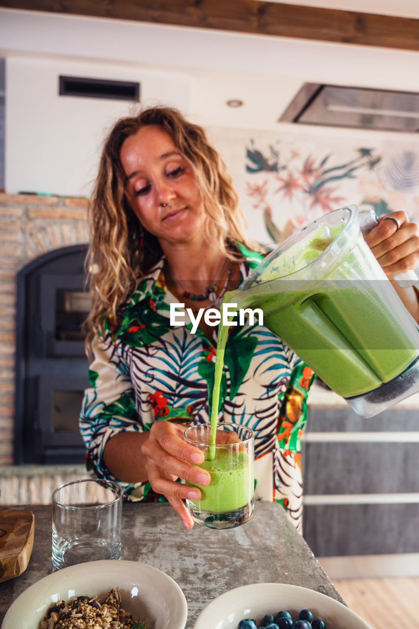 Attractive female pouring healthy green smoothie from blender bowl into glass while standing near table in light kitchen at home
