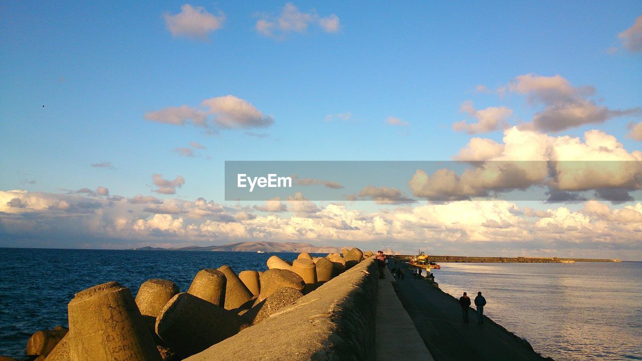 People walking on pier by groyne rocks in sea against sky