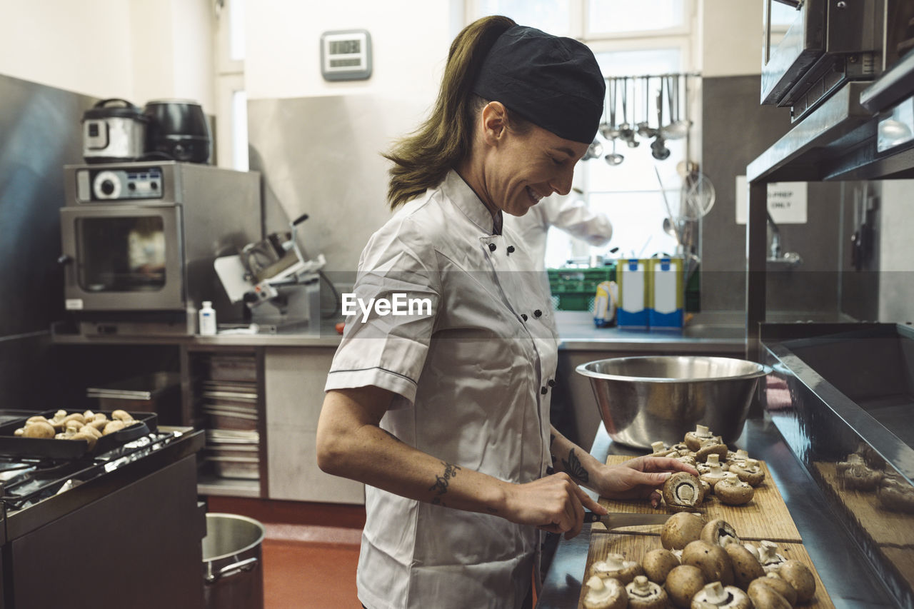 Side view of smiling female chef cutting mushrooms at kitchen counter