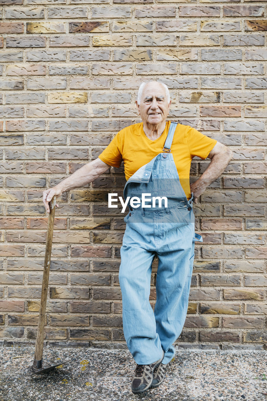 Senior man in denim overall with garden tool looking at camera leaning on a wall